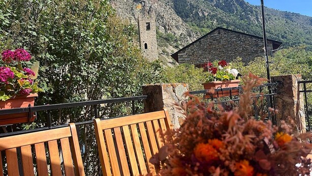 A terrace with wooden furniture, potted flowers, and a stone building with a tower in the background. - Country House with stunning views near Porte-Puymorens and Andorra (Carol)