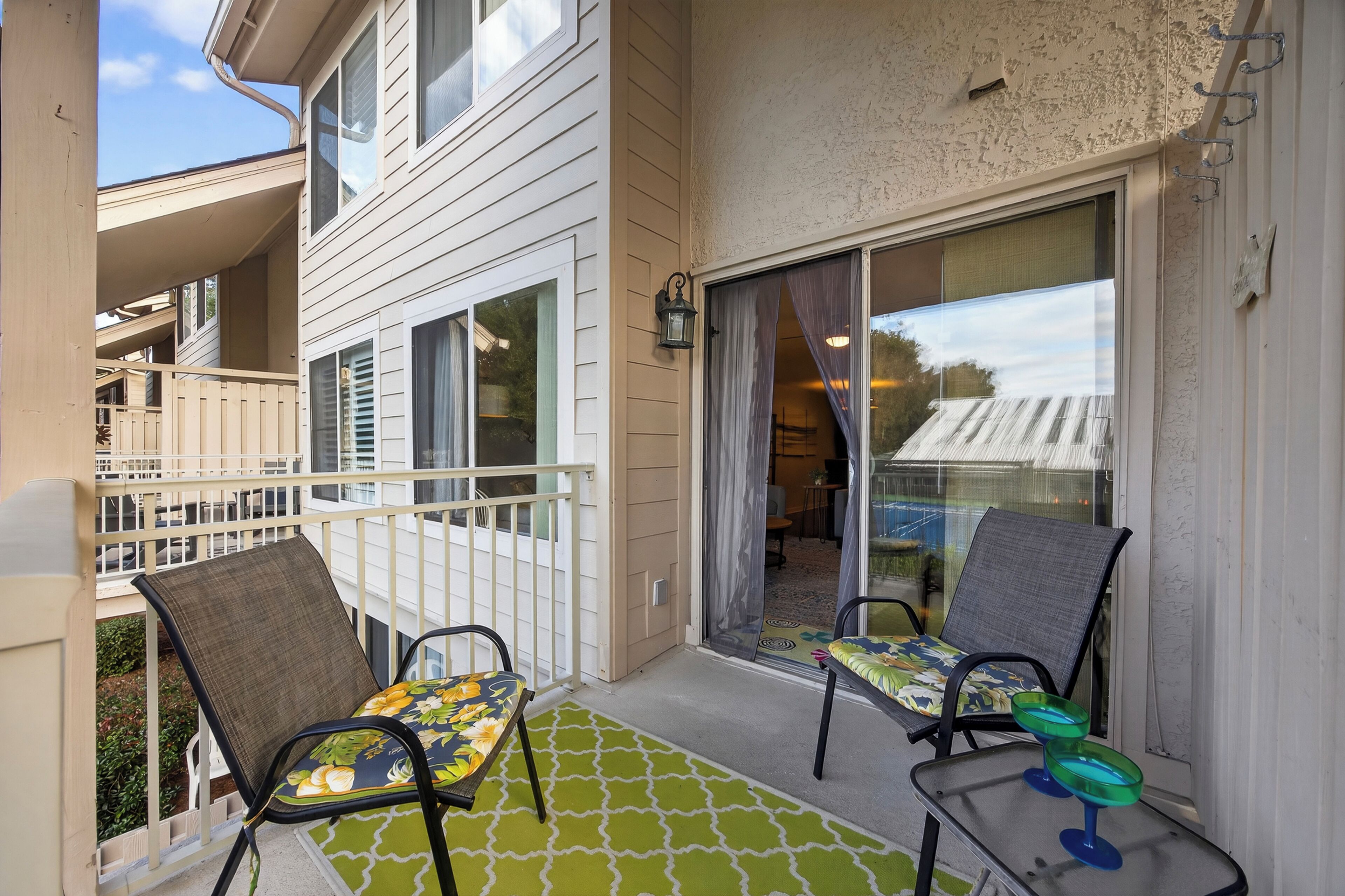 A balcony with two chairs and a table, a sliding glass door, and a small outdoor area with a patterned rug.