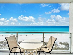 Ein Balkon mit Tisch und Stühlen mit Blick auf einen Strand, Menschen und einen klaren Himmel.