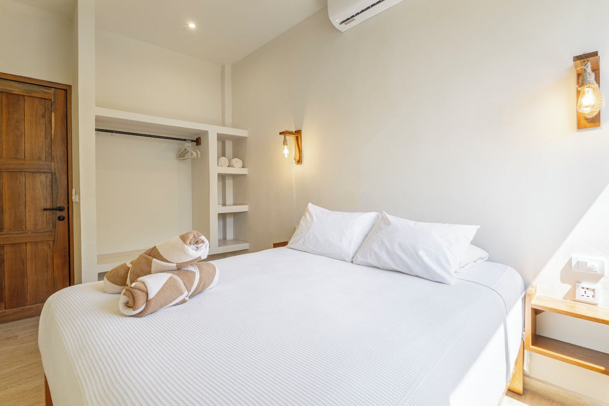 A neatly made bed with white linens and a brown towel, a wooden door, and a wall-mounted light fixture.