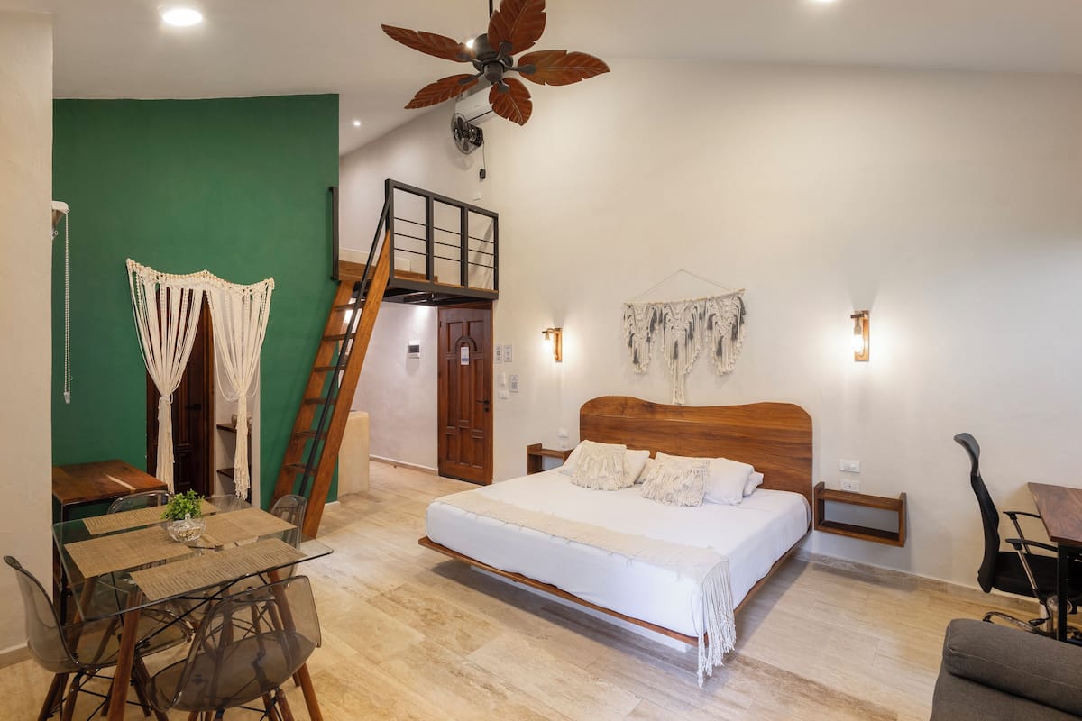 A bedroom with a wooden bed, a dining table with chairs, a staircase, and a green wall.