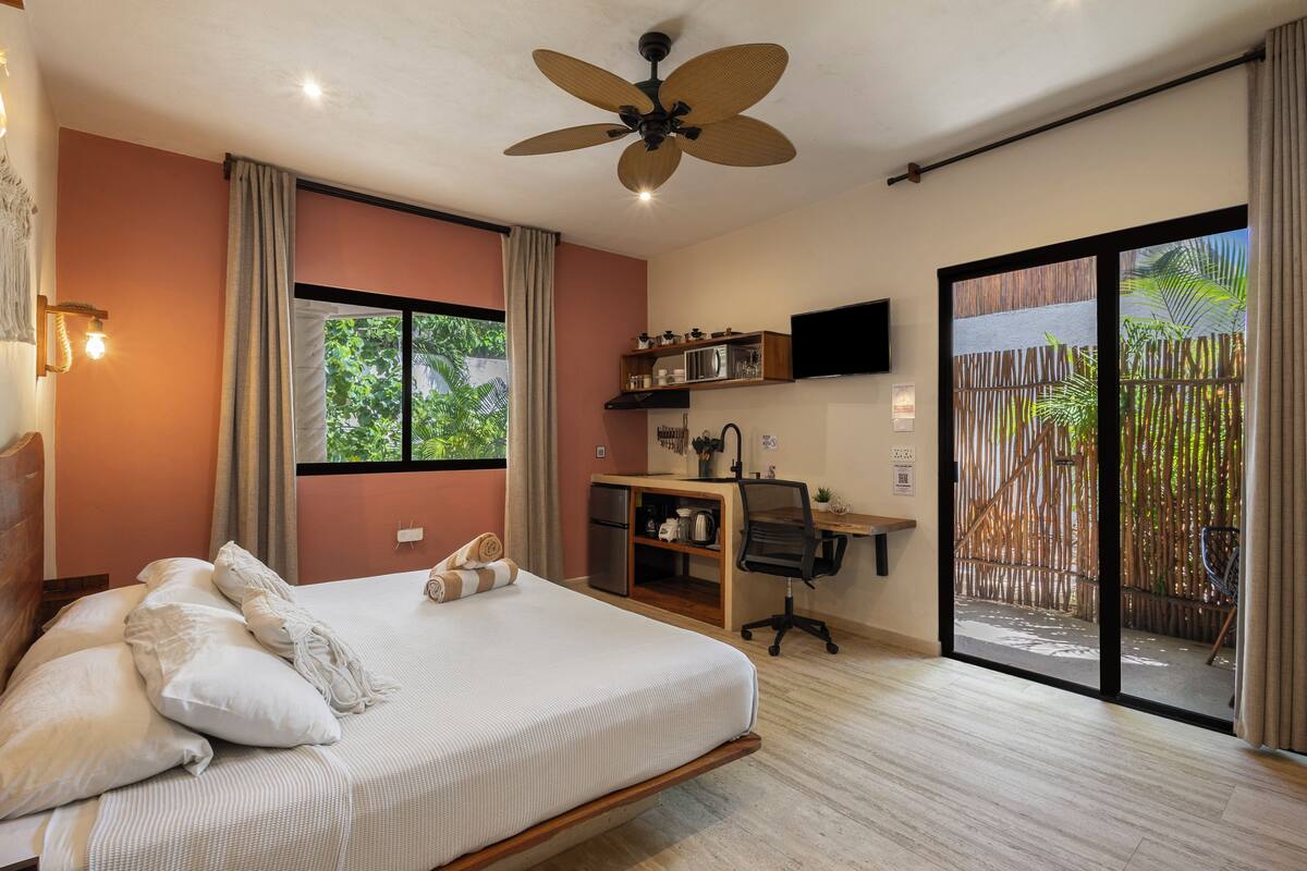 A bedroom with a bed, a desk, a chair, a ceiling fan, and a sliding glass door to a balcony.