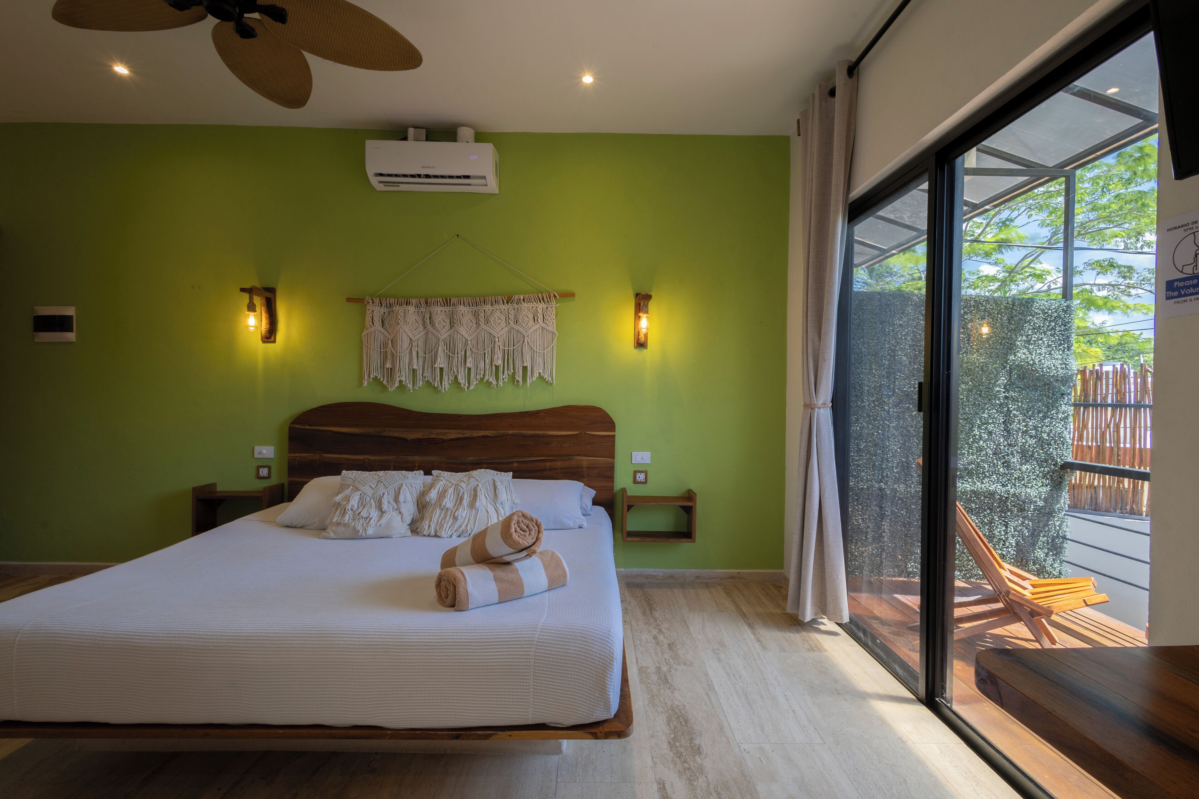 Un dormitorio con una cama, un ventilador de techo, un equipo de aire acondicionado, luces empotradas en la pared, un tapiz de macramé y vistas al exterior a través de una puerta corrediza de vidrio.