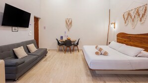 A hotel room with a grey sofa, a wooden bed, a desk with two chairs, a flat-screen TV mounted on the wall, and a wall-mounted light fixture. - Casa Wayra Tulum - Boutique Condos (Tulum)