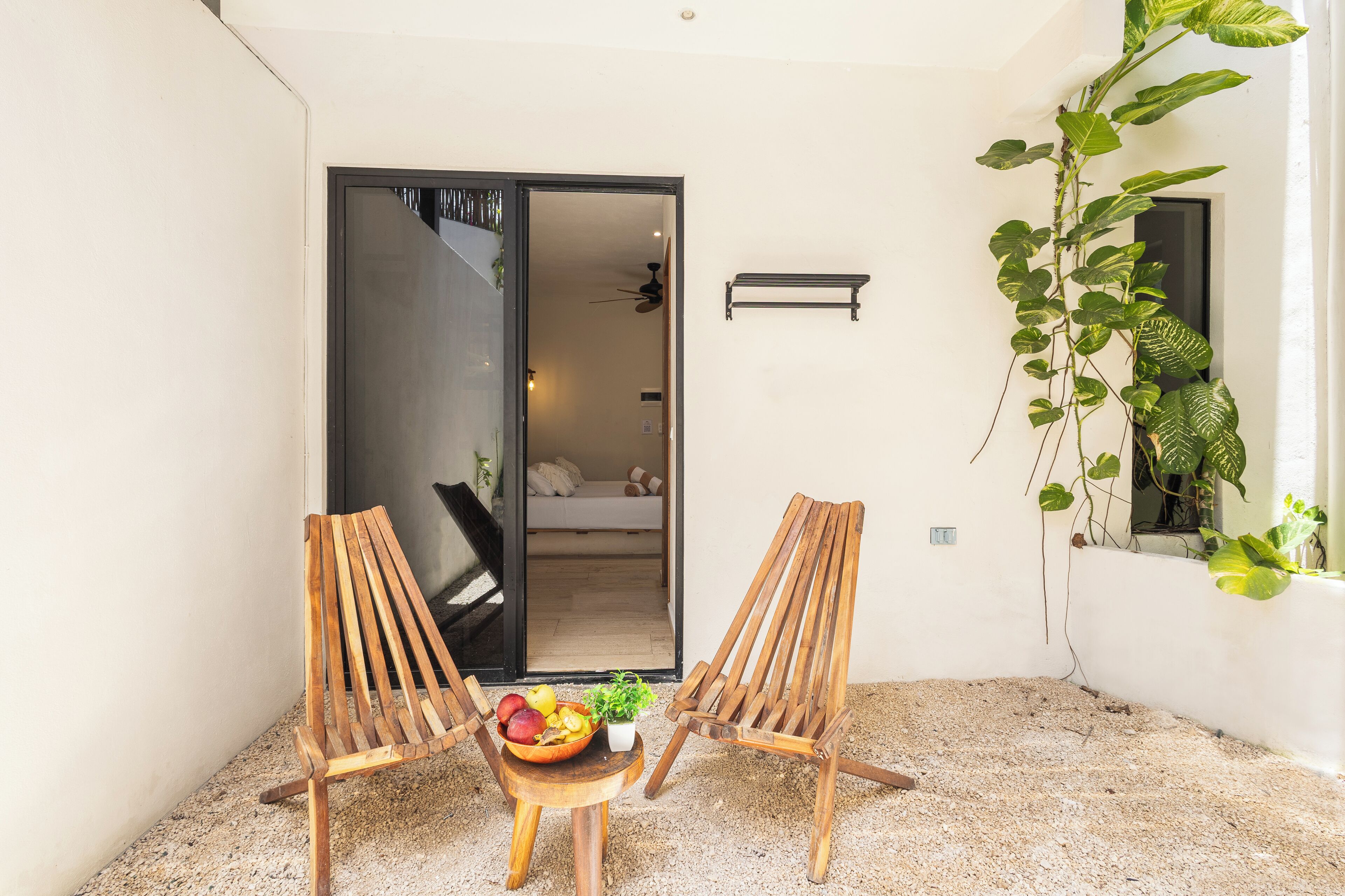 A patio with two wooden chairs, a small table with fruit and a plant, and a sliding glass door leading to a bedroom.