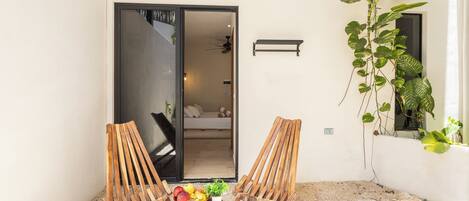 A patio with two wooden chairs, a small table with fruit and a plant, and a sliding glass door leading to a bedroom.