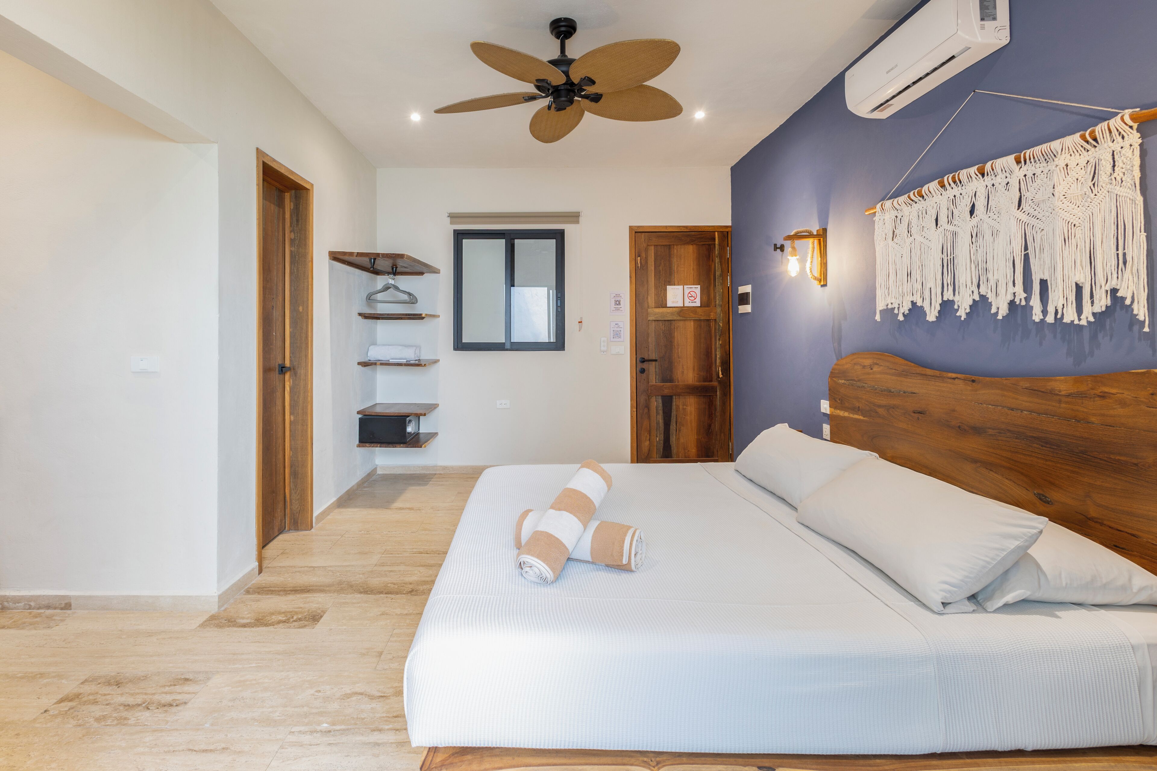 Un dormitorio con cabecera de madera, ventilador de techo, aire acondicionado de pared, un tapiz de macramé y cama con ropa de cama blanca.