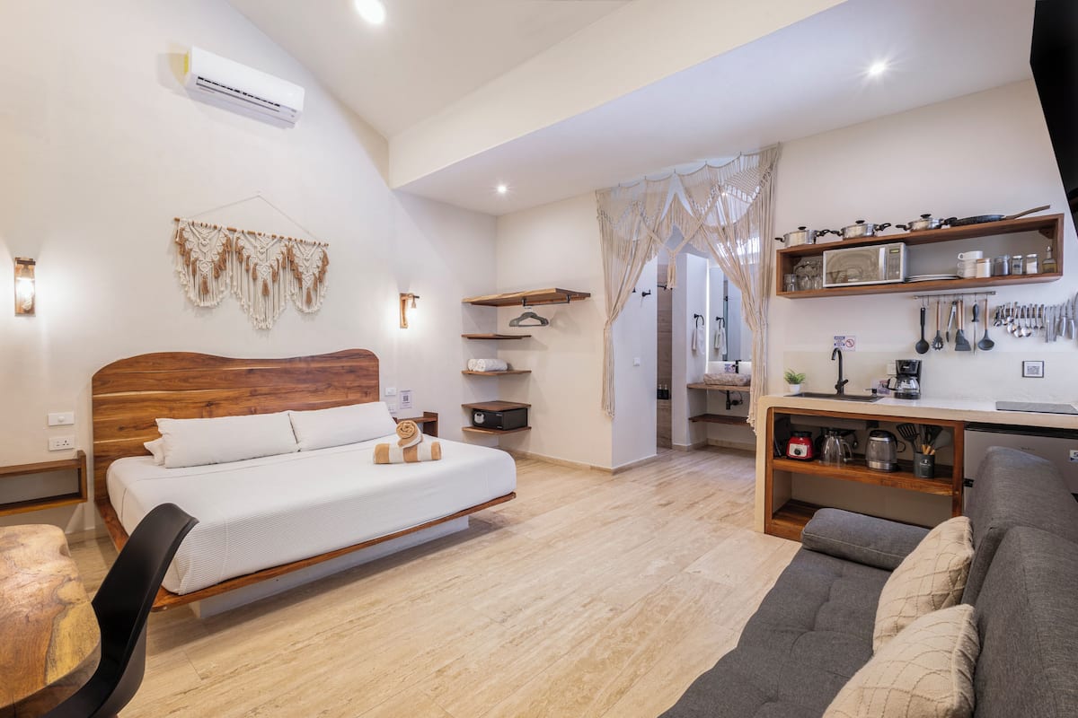 A modern bedroom with a wooden bed, bedside tables, a grey sofa, and a kitchenette area with a microwave and utensils.