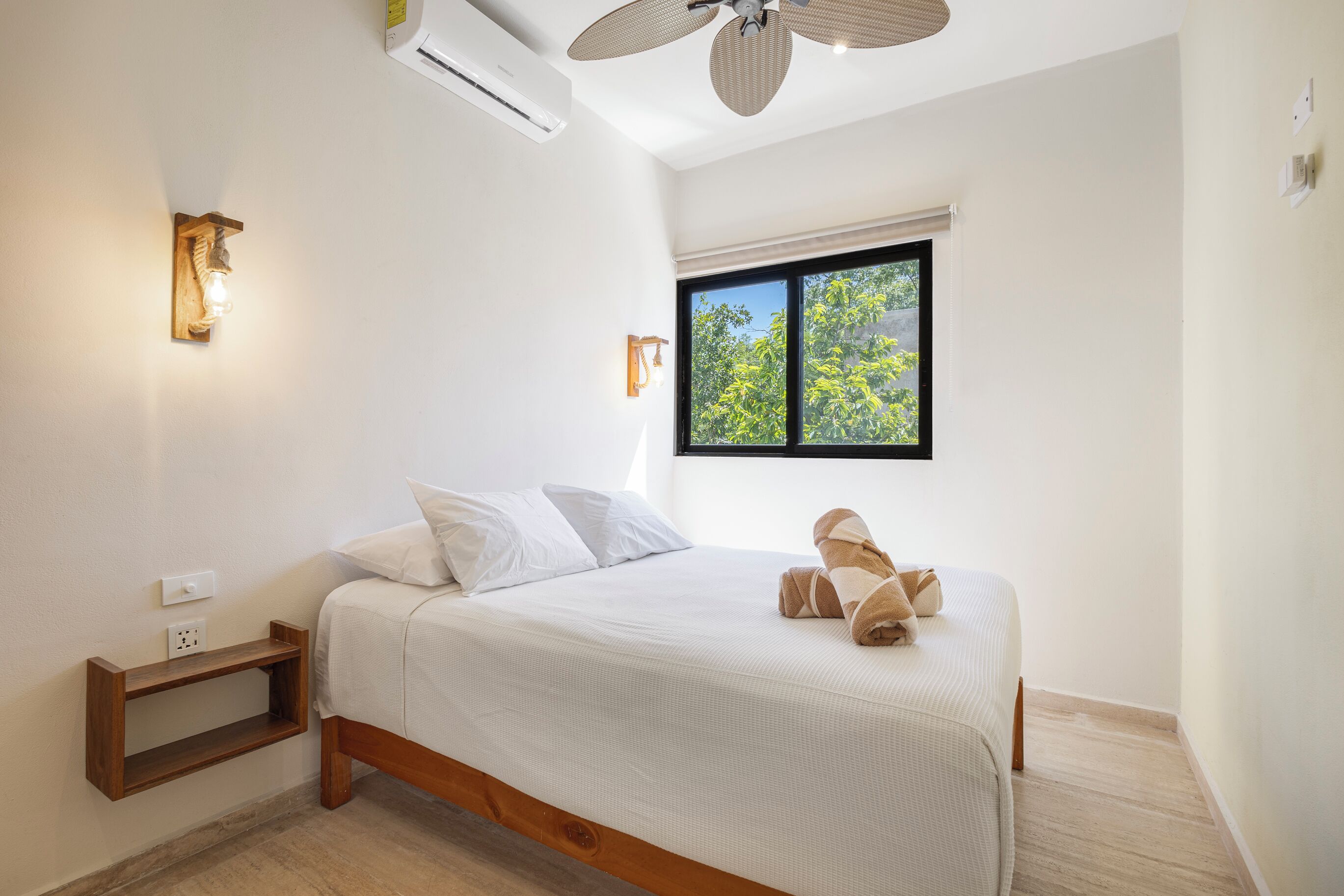 A bedroom with a bed, bedside table, wall-mounted light, window, and ceiling fan.