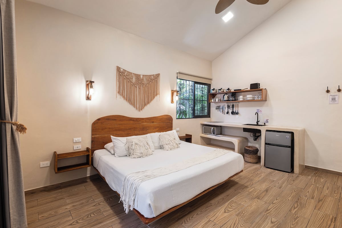 A bedroom with a wooden bed, bedside tables, a small kitchenette, and a window with a view of greenery.