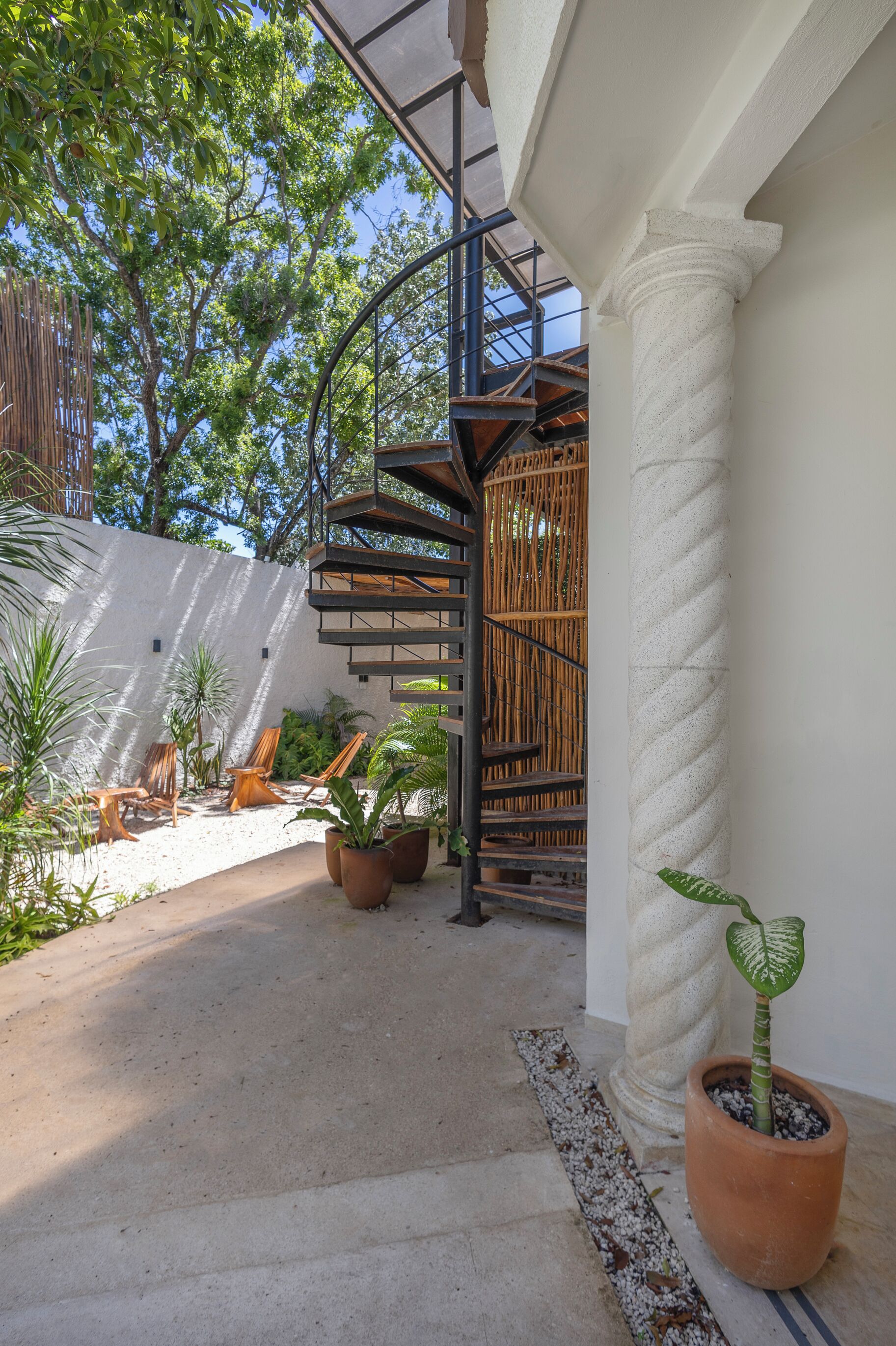 Un espacio exterior moderno con una escalera en espiral, sillas de madera y plantas en macetas.