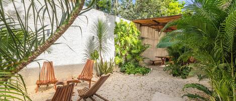 A patio area with wooden chairs and a table, surrounded by tropical plants and a white wall.