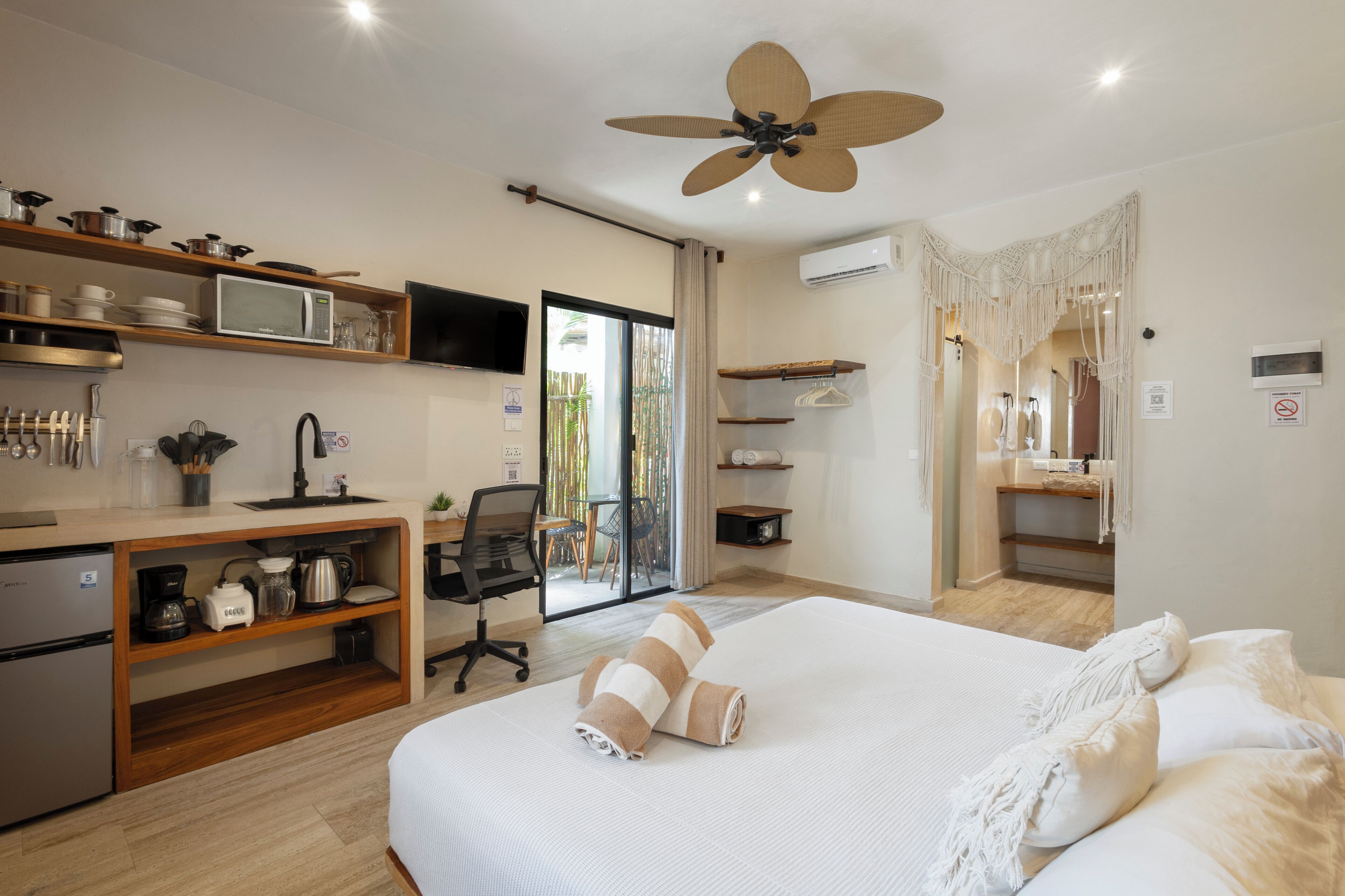 A modern hotel room with a large bed, a desk with a computer, a kitchenette, and a sliding glass door to the balcony.