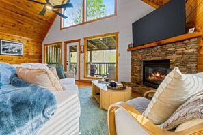 Ein gemütliches Blockhaus-Interieur mit Kamin aus Naturstein, Holzwänden und einem großen Fenster mit Blick auf eine Terrasse.