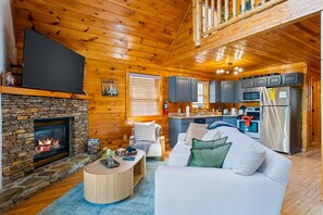 Ein gemütliches Blockhaus-Interieur mit Kamin aus Naturstein, einem Wohnzimmer mit Sofa und Couchtisch sowie einer Küche mit modernen Geräten und Holzbalkendecken.