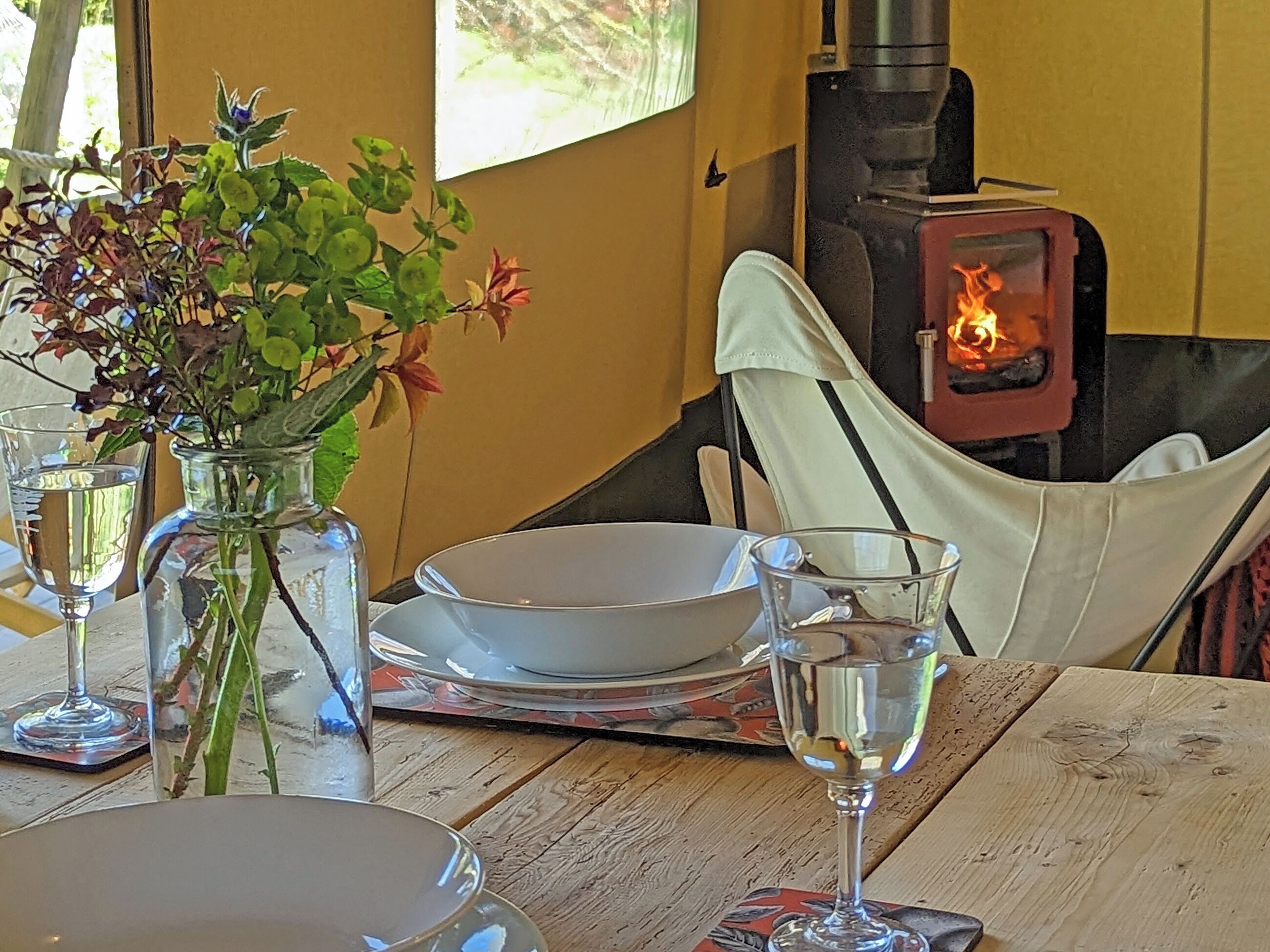 A dining table with plates, glasses, and a vase with flowers. A wood-burning stove is visible in the background.