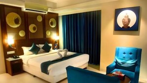 A hotel room with a bed, a blue armchair, a small table, and a wall-mounted air conditioner. - Annex Suites Bogura (Bogra)