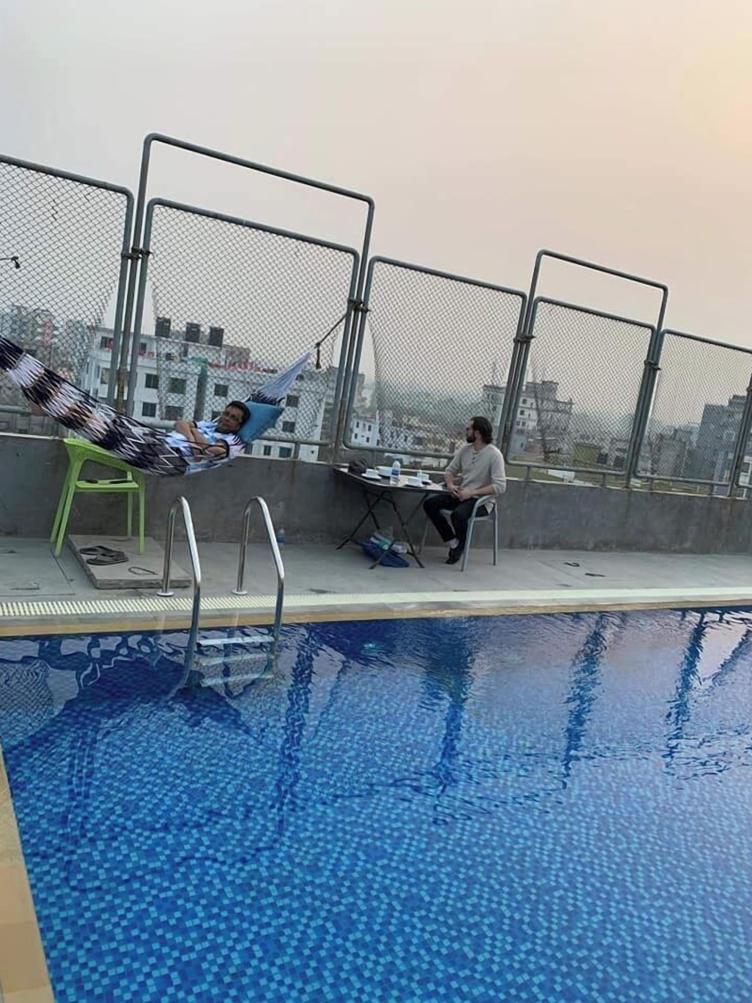 Una piscina en la azotea con una persona descansando en una hamaca y otra sentada a una mesa.