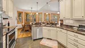 Una cocina con gabinetes blancos, encimeras de granito, lavavajillas y un comedor con mesa y sillas de madera.