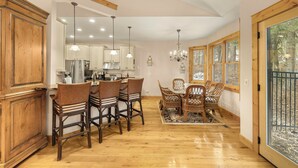 Una cocina amplia con armarios de madera, un comedor con sillas de mimbre y ventanas grandes con vistas a árboles.