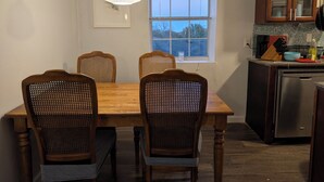 Een eethoek met een houten tafel en vier stoelen, een hangende lamp, een raam met uitzicht op bomen, en een keuken met kasten en een vaatwasser.