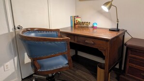 Een houten bureau met een lamp, een blauwe stoel met metalen details, een klein decoratief object en een lijst met een foto aan de muur.