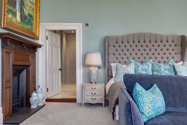 A bedroom with a tufted headboard, a bedside table with a lamp, a dresser, and a fireplace.