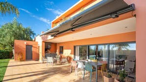 Een modern overdekt terras met eethoek, een barbecue en grote schuiframen naar een tuin.