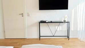 Une chambre à coucher avec un lit, un téléviseur fixé au mur, une table de chevet avec un livre et un vase, et une porte.