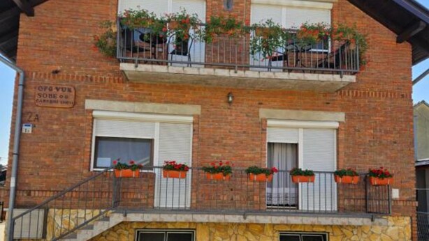 Un immeuble à deux étages, construit en briques, avec un balcon au dernier étage, agrémenté de jardinières et d’un escalier menant à l’entrée.