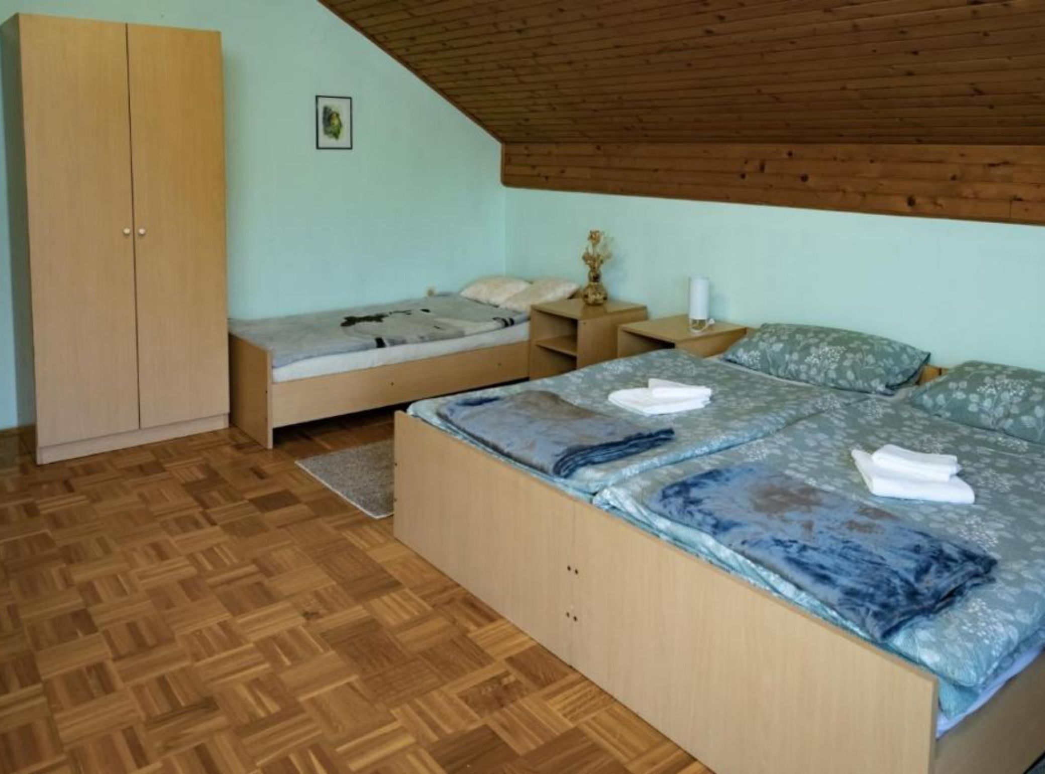 A bedroom with a wooden wardrobe, a bed with a floral duvet, a nightstand with a lamp, and a wooden floor.