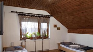 Une chambre mansardée chaleureuse avec un plafond en pente, deux lits, une fenêtre ornée de rideaux légers et des plantes en pot sur le rebord de la fenêtre.