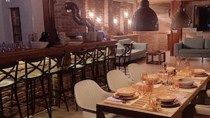 A dining area with a long wooden table set for a meal, surrounded by chairs. There is a bar area with stools to the left and a sofa in the background.
