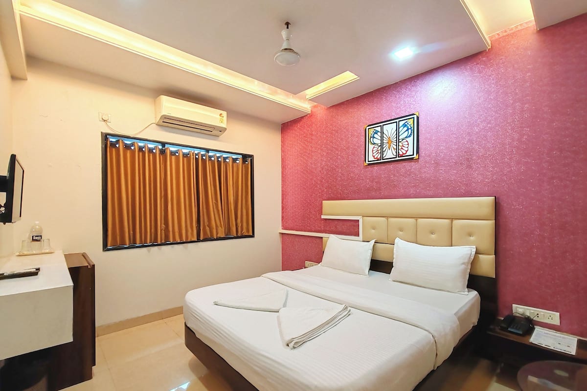 A hotel room with a bed, a desk, an air conditioner, a window with curtains, and a decorative piece on the wall.