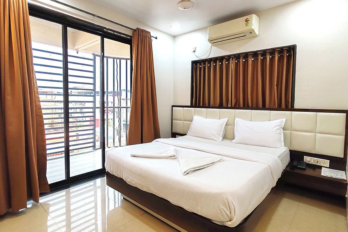 A hotel room with a bed, curtains, an air conditioning unit, and a view through a window.