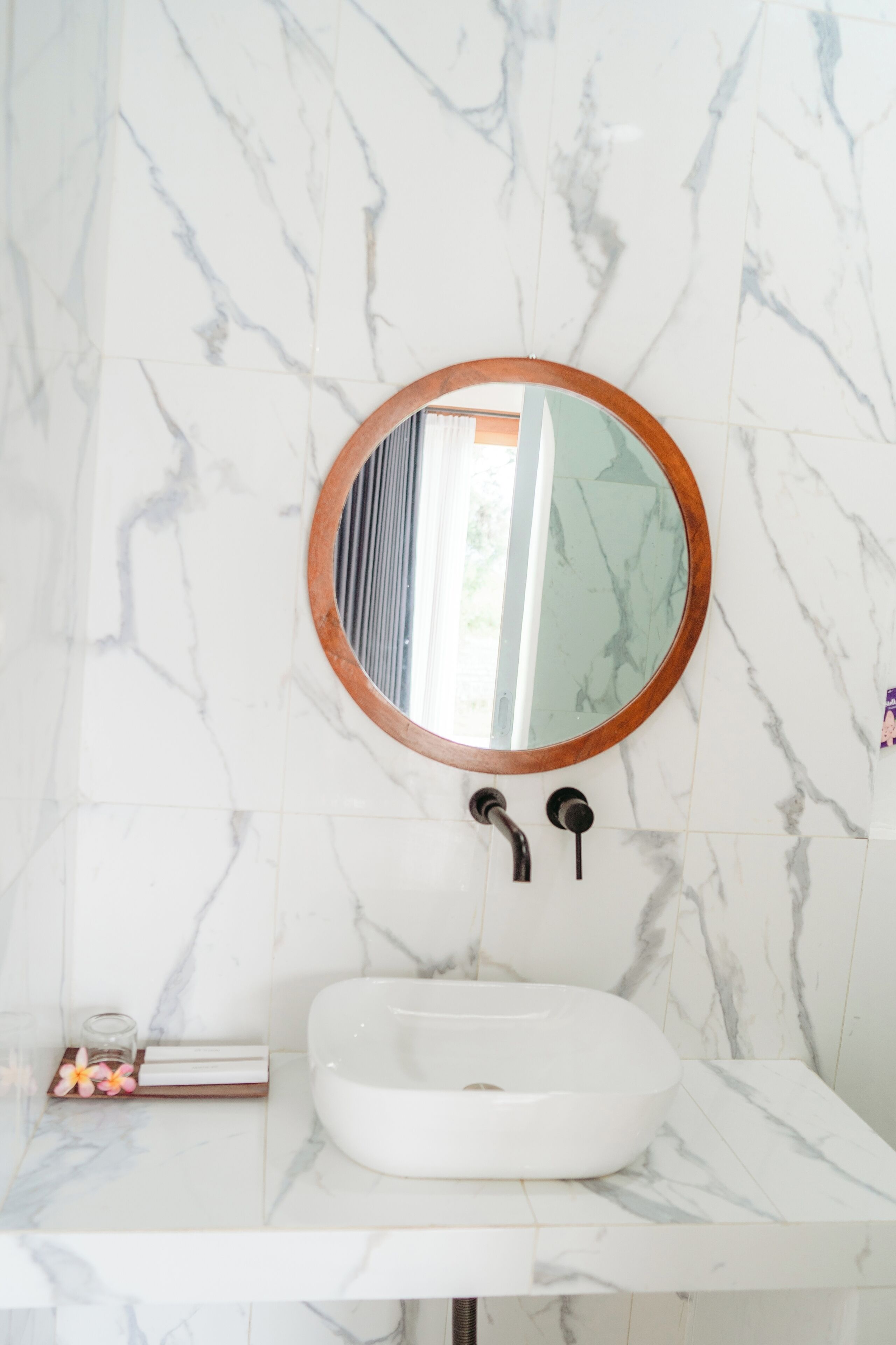 A bathroom with a marble wall, a round mirror with a wooden frame, a white sink, and a small glass container with flowers.