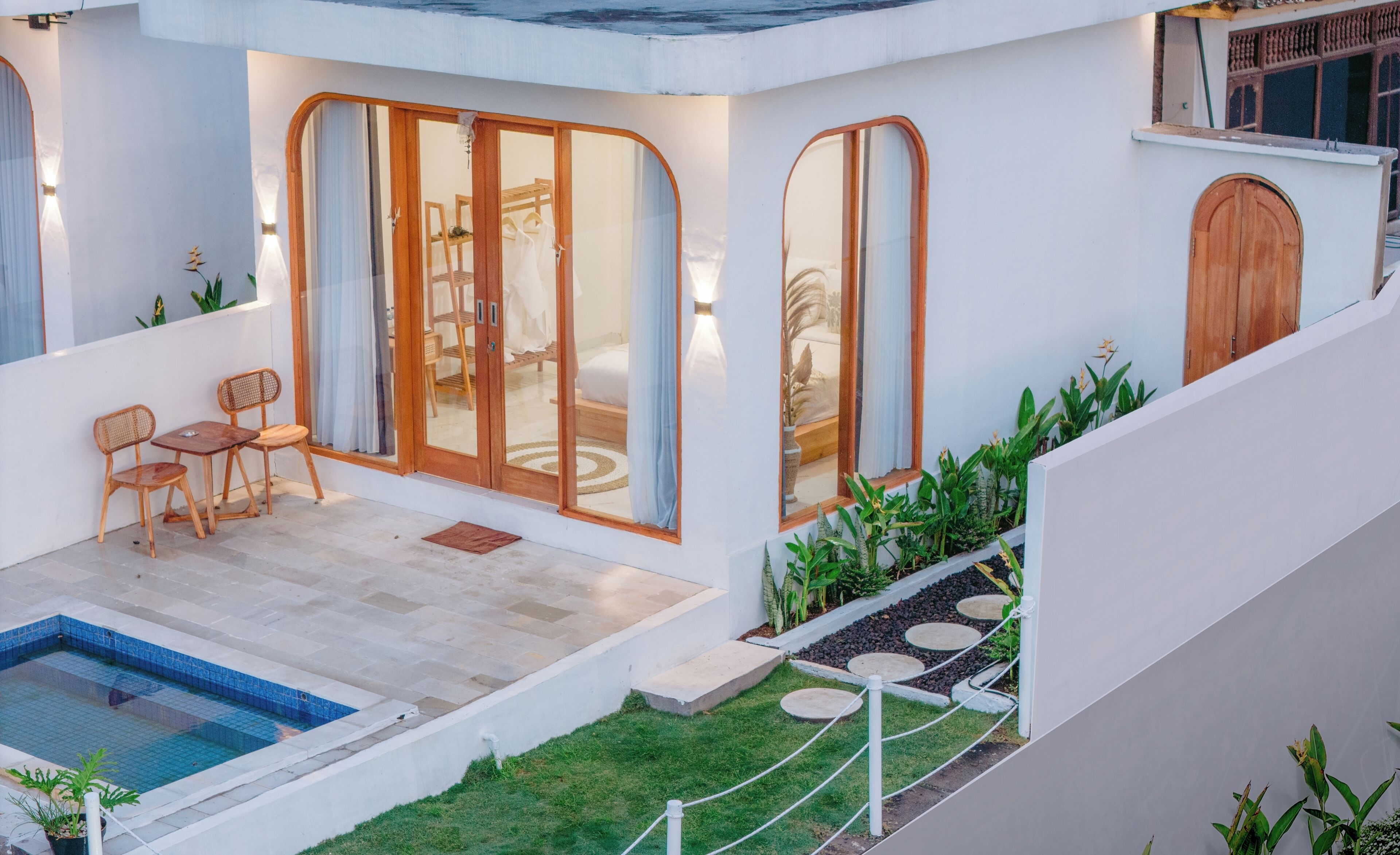 A modern house with a swimming pool, a dining area with wooden furniture, and a garden with plants and stepping stones.
