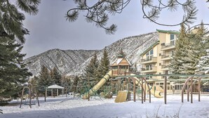 Une aire de jeux recouverte de neige, avec des toboggans et des balançoires, entourée d’arbres et bordée d’un bâtiment doté de balcons.