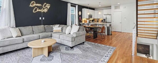 En moderne stue med en grå sofa, et rundt sofabord og et tæppe. Der er et køkkenområde med hvide skabe og et spisebord.
