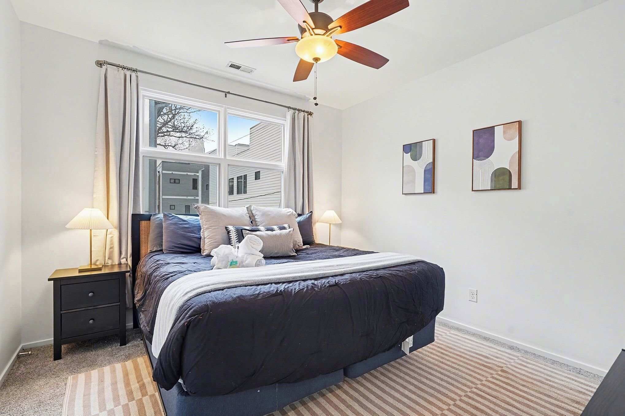 A bedroom with a bed, two bedside lamps, a ceiling fan, and two abstract wall art pieces.