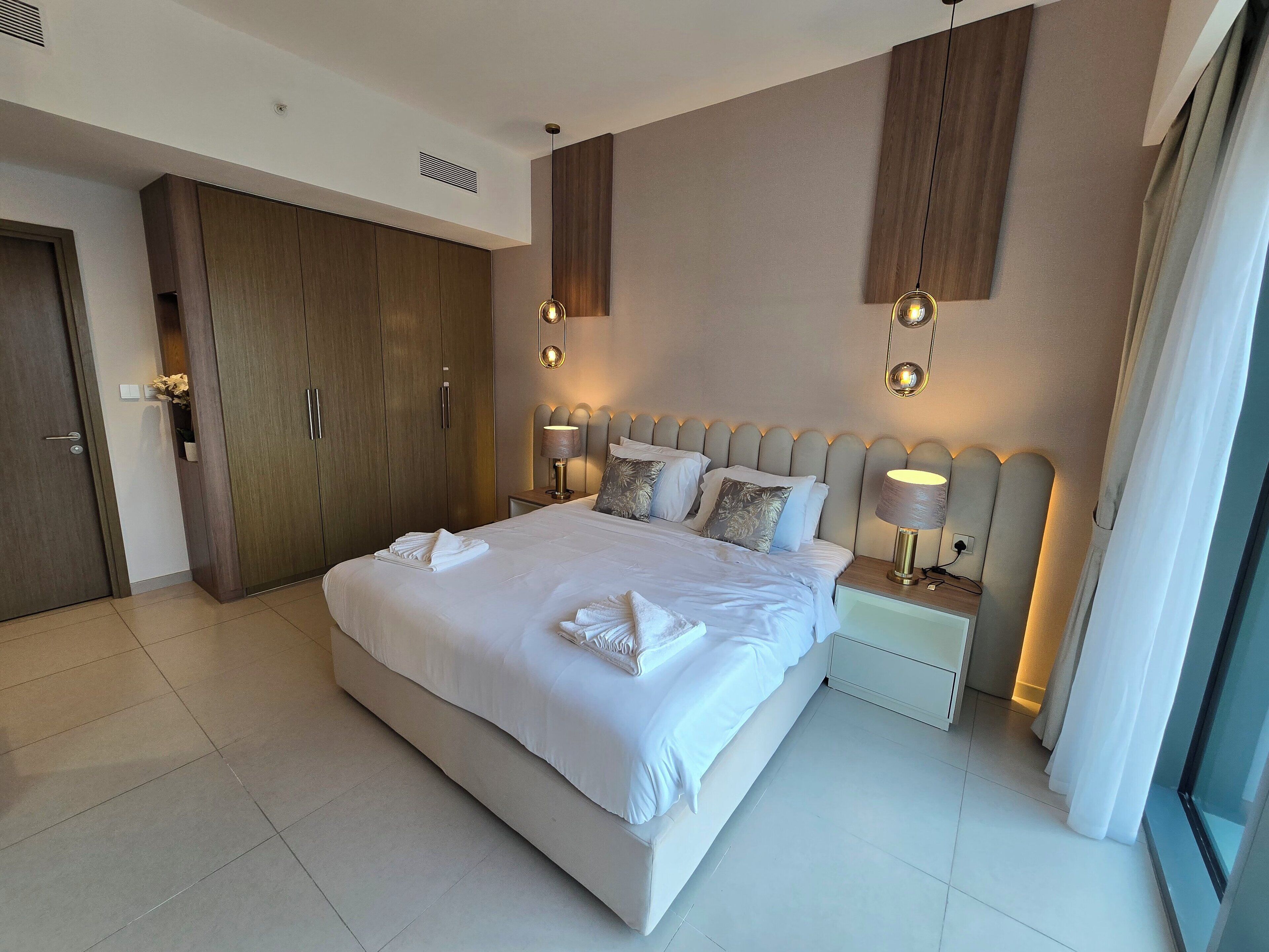 A modern hotel room with a large bed, two bedside tables, a wardrobe, and a large window.