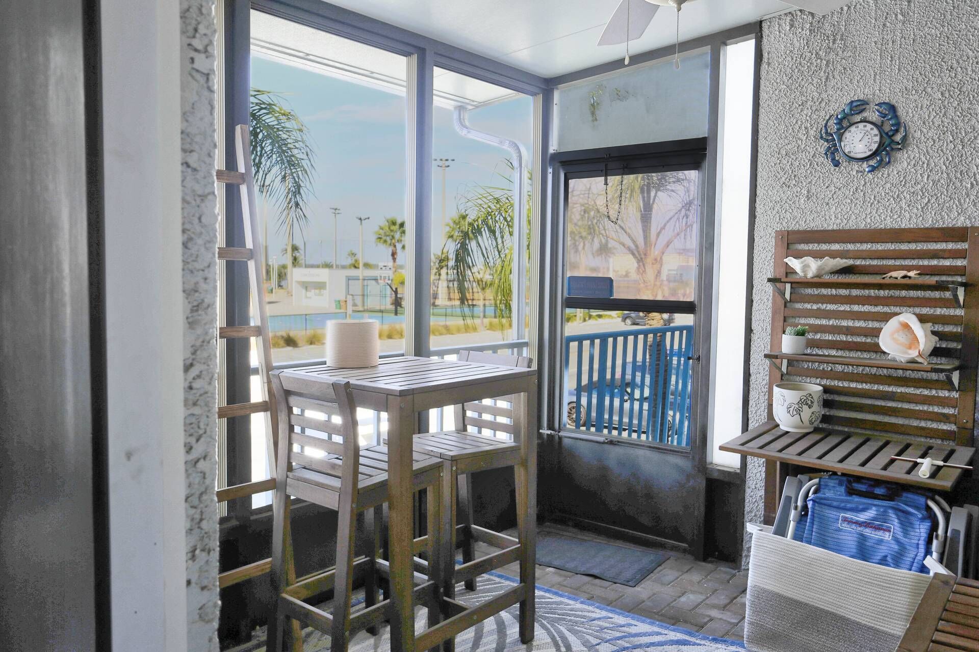 A room with a wooden table and chairs, a view of the outdoors, a balcony with a railing, and a wall-mounted shelf with decorative items.