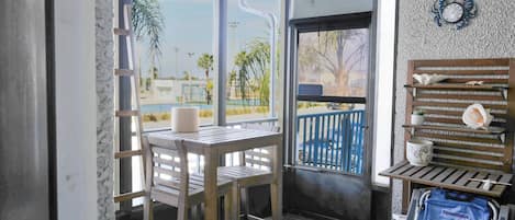 A room with a wooden table and chairs, a view of the outdoors, a balcony with a railing, and a wall-mounted shelf with decorative items.