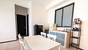 Eine kleine Küche mit weißem Tisch und Stühlen, einem schwarzen Kühlschrank, einer Kaffeemaschine und einem Fenster mit Jalousien.