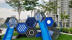 A playground with slides and climbing structures, surrounded by green grass and trees, with high-rise buildings in the background.
