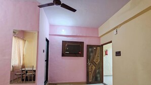 A room with pink walls, a ceiling fan, a TV mounted on the wall, a door with a number, and a bed with a red cover. - Hotel Pallavi Inn (Panvel)