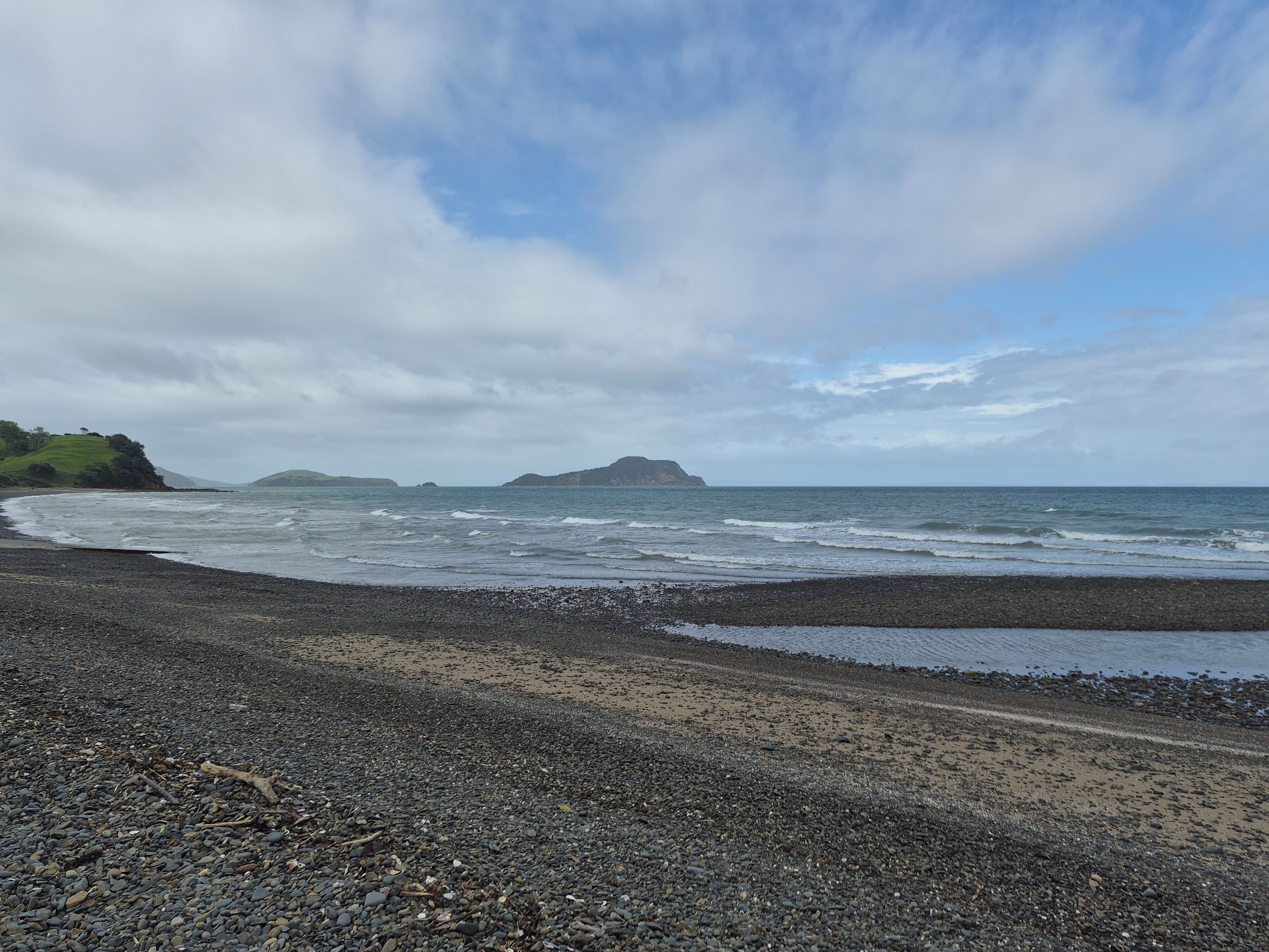 Une plage de galets, des vagues, une île lointaine et un ciel nuageux.