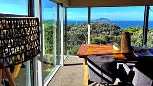 A balcony with a wooden table and a cushioned bench, overlooking a lush landscape and the sea.