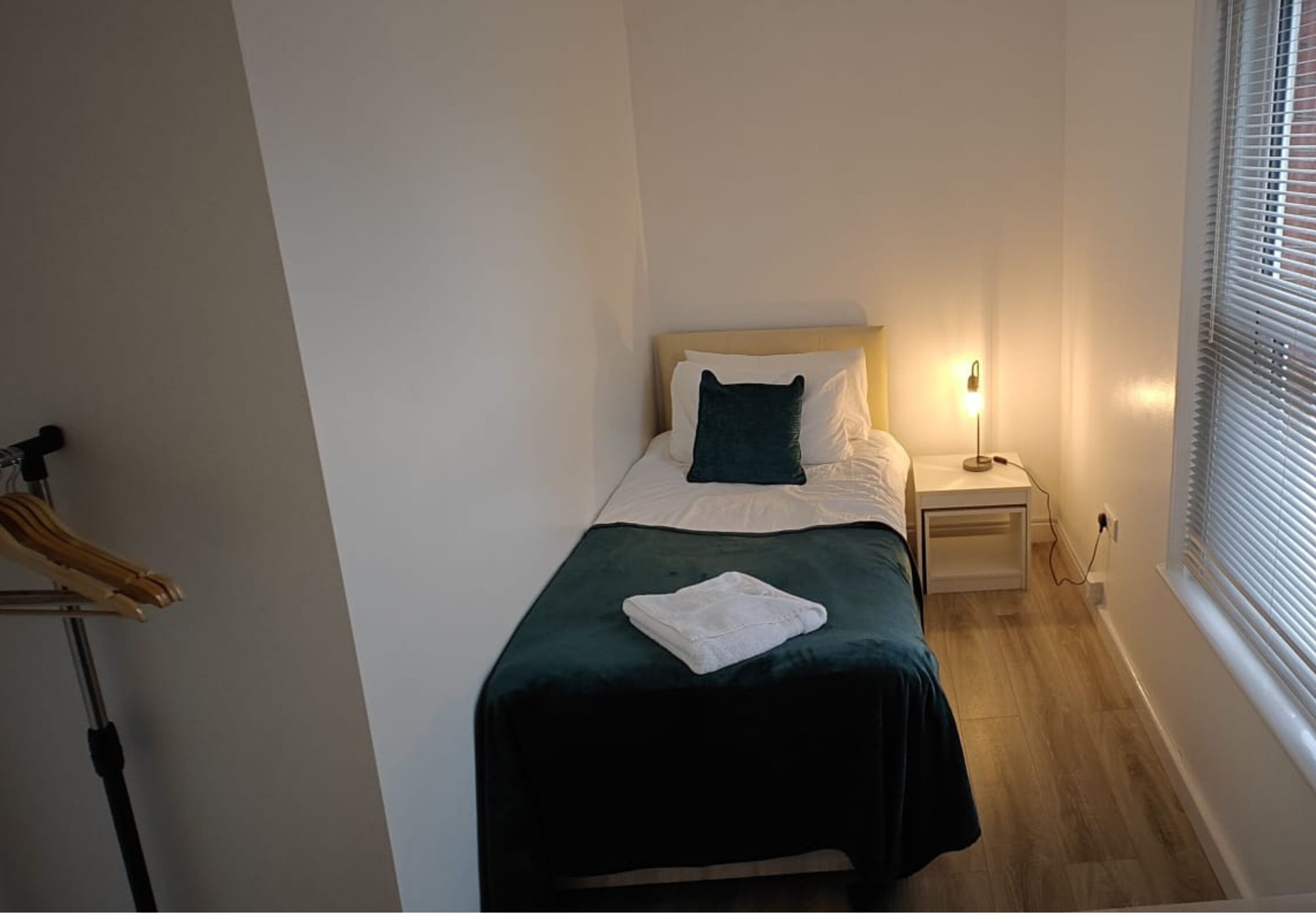 A small, neatly arranged bedroom with a bed, bedside lamp, and a window with blinds.