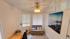 Una habitación con un escritorio de madera, una silla de oficina negra, una cama con almohadas, un ventilador de techo y una ventana grande con persianas.